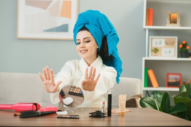 Jovem sorridente enrolada em uma toalha de cabelo com unhas de gel secas, sentada à mesa com ferramentas de maquiagem na sala de estar