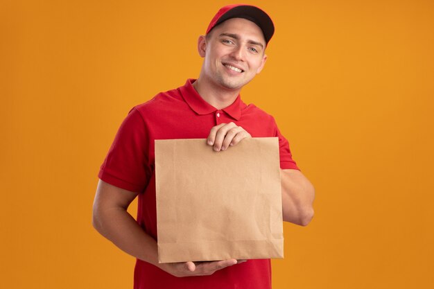 Jovem sorridente entregador de uniforme com tampa segurando um pacote de comida de papel isolado na parede laranja