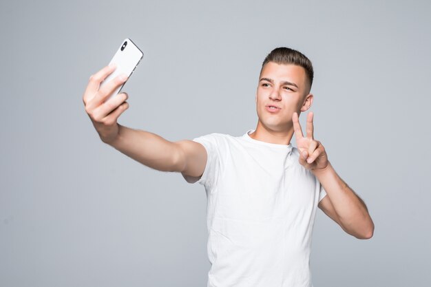 Jovem sorridente está vestindo uma camiseta branca e fazendo um sinal de vitória de selfie com um smartphone prata.