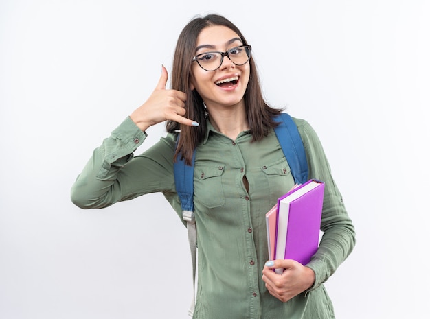 Jovem sorridente, estudante, usando óculos e mochila segurando livros, mostrando um gesto de ligação