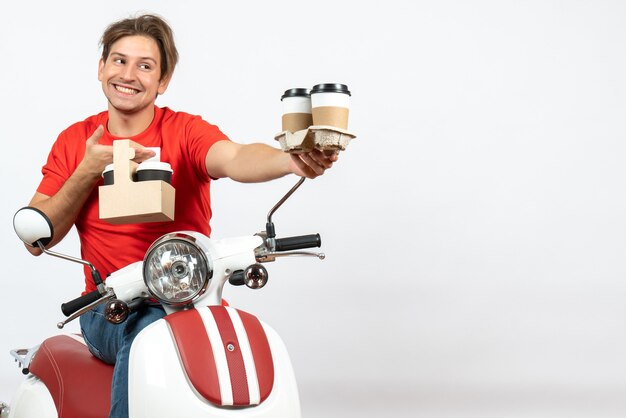 Jovem sorridente feliz mensageiro de uniforme vermelho sentado na scooter segurando pedidos na parede amarela