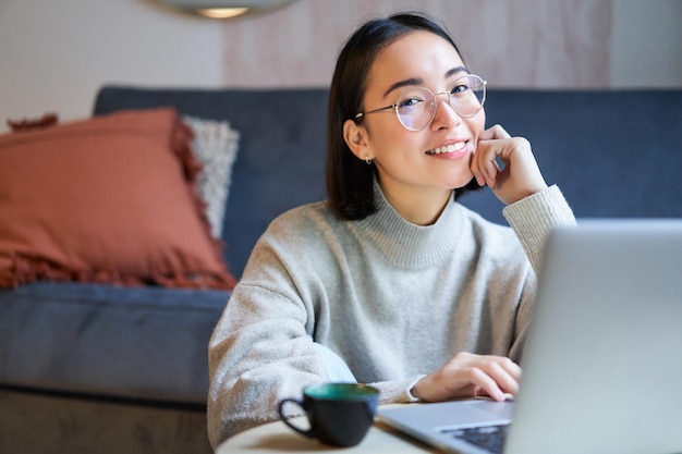 Jovem sorridente freelancer autônoma que fica em casa trabalhando no controle remoto do laptop usando gl