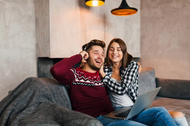 Jovem sorridente homem e mulher sentados em casa no inverno segurando no laptop ouvindo fones de ouvido com expressão de rosto chocado e surpreso, usando a internet, casal em momentos de lazer juntos, felizes