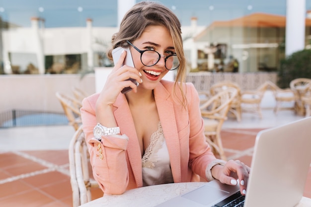 Jovem sorridente, linda mulher de negócios, usando óculos elegantes, jaqueta rosa inteligente, relógios brancos. Falando por telefone, trabalhando em um laptop prateado do lado de fora em um café.