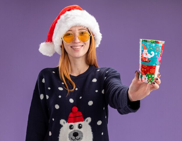 Jovem sorridente linda vestindo suéter de natal e chapéu com óculos segurando uma taça de natal isolada na parede roxa