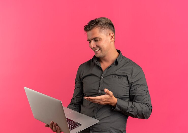Foto grátis jovem sorridente loira bonita segura e aponta para um laptop isolado em um espaço rosa com espaço de cópia