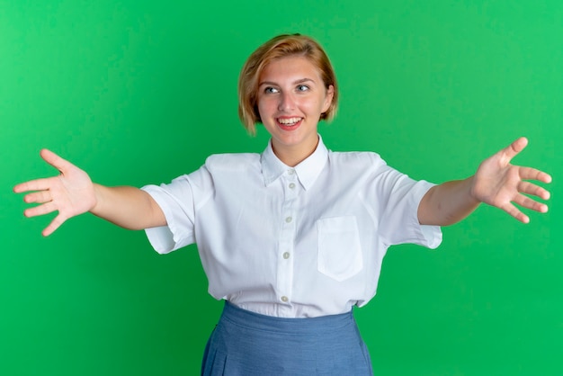 Foto grátis jovem sorridente loira russa de braços abertos, olhando para o lado isolado em um fundo verde com espaço de cópia