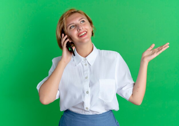Jovem sorridente loira russa falando ao telefone com a mão levantada isolada em um fundo verde com espaço de cópia
