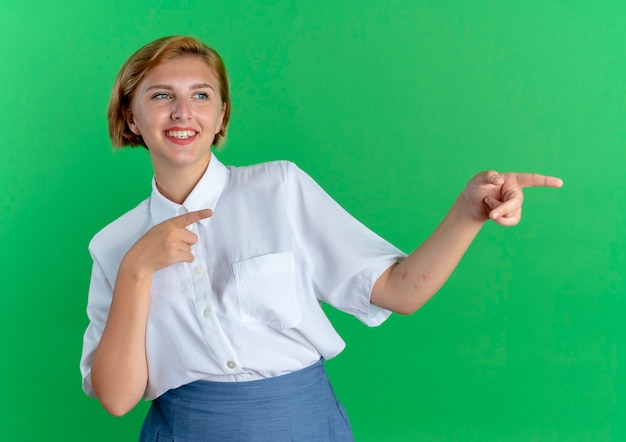 Jovem sorridente loira russa olha e aponta para o lado isolado em um fundo verde com espaço de cópia