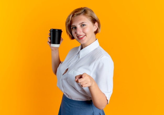 jovem sorridente loira russa parada de lado segurando uma xícara de café e apontando para a câmera