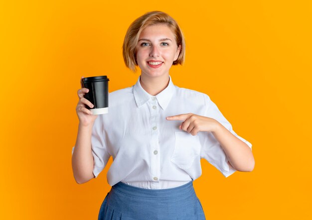 Jovem sorridente loira russa segura e aponta para a xícara de café isolada em um fundo laranja com espaço de cópia