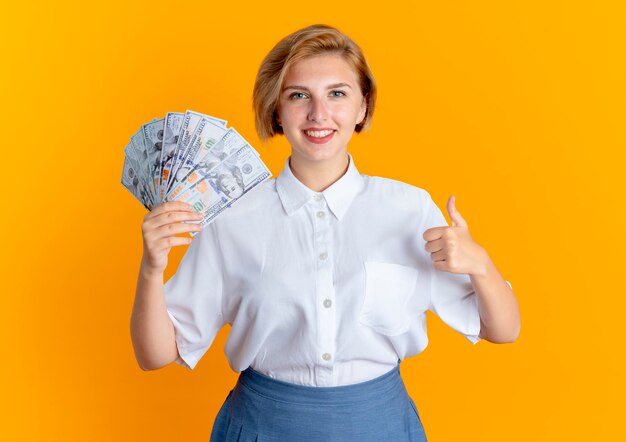 Jovem sorridente loira russa segurando dinheiro e polegares para cima, isolados em um fundo laranja com espaço de cópia