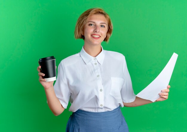 Jovem sorridente loira russa segurando uma xícara de café e folhas de papel isoladas em um fundo verde com espaço de cópia