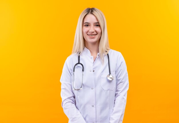 Jovem sorridente médico usando estetoscópio em bata médica e aparelho dentário em fundo amarelo isolado