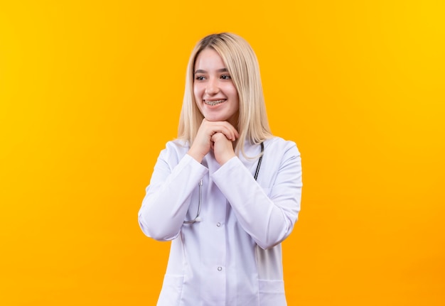 Jovem sorridente, médico, usando estetoscópio, vestido de médico e aparelho dentário, colocando as duas mãos sob o queixo em fundo amarelo isolado