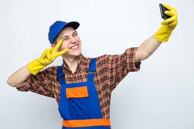 Jovem sorridente, mostrando gesto de paz, faxineiro usando uniforme e boné com luvas tirando uma selfie