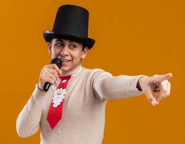 Jovem sorridente olhando de lado usando chapéu com gravata natalina falando nos pontos do microfone ao lado isolado na parede amarela
