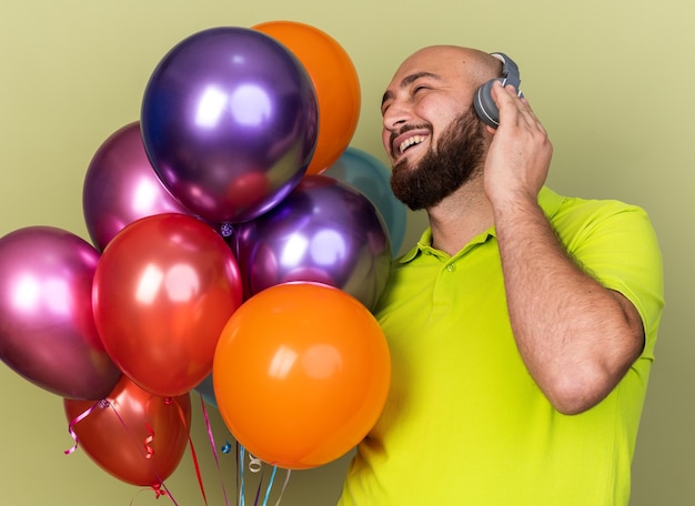 Foto grátis jovem sorridente, olhando para o lado, vestindo camiseta amarela e fones de ouvido segurando balões isolados na parede verde oliva