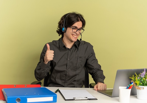 Jovem sorridente trabalhador de escritório com fones de ouvido em óculos ópticos sentado na mesa com ferramentas de escritório usando e olhando para o laptop, polegares para cima