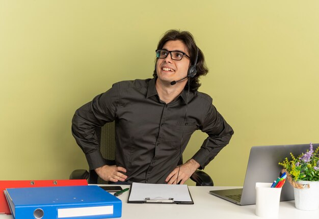 Jovem sorridente trabalhador de escritório com fones de ouvido em óculos ópticos, sentado na mesa com ferramentas de escritório, usando laptop e olhando para o lado isolado em um fundo verde com espaço de cópia