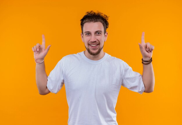 Jovem sorridente vestindo uma camiseta branca apontando para cima em um fundo laranja isolado