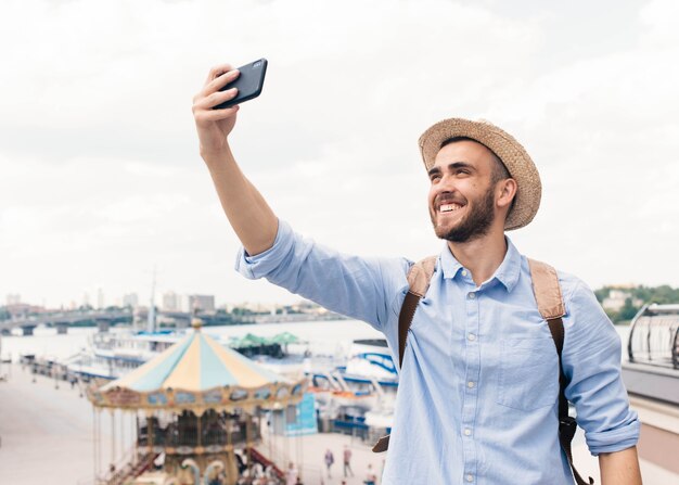 Jovem, sorrindo, homem, segurando, telefone pilha, e, levando, selfie, ao ar livre