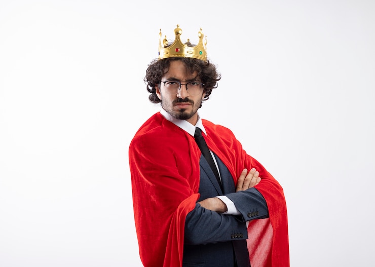 Foto grátis jovem super-herói caucasiano sério usando óculos óticos, usando um terno com capa vermelha e a coroa está de lado com os braços cruzados