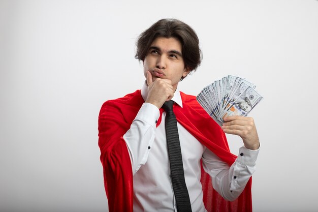 Foto grátis jovem super-herói pensando olhando para o lado usando gravata, segurando dinheiro e segurando o queixo isolado no branco