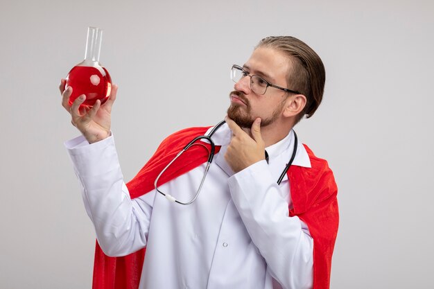 Jovem super-herói pensando vestindo túnica médica com estetoscópio e óculos segurando e olhando para a garrafa de vidro de química cheia de líquido vermelho, colocando a mão no queixo