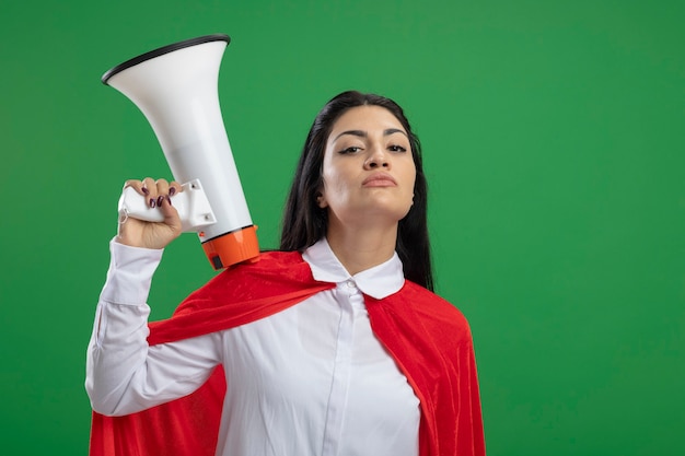 Jovem super-heroína caucasiana presunçosa segurando um alto-falante na mão e exibindo expressões faciais confiantes isoladas na parede verde com espaço de cópia