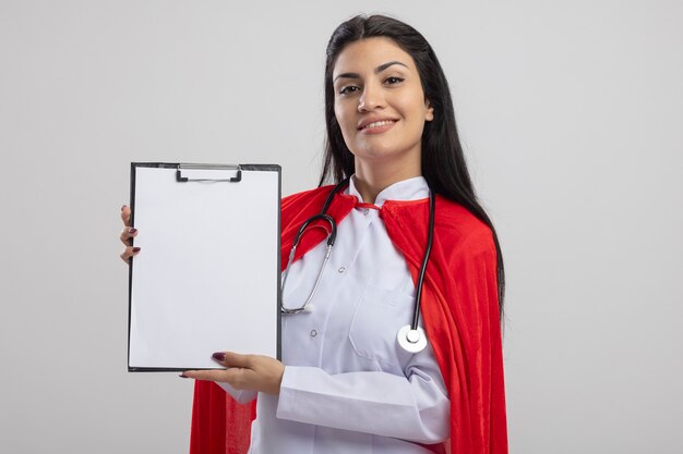 Jovem super-heroína caucasiana sorridente com estetoscópio mostrando a prancheta, olhando para a câmera isolada no fundo branco com espaço de cópia