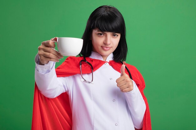 Jovem super-heroína confiante usando um estetoscópio com um manto médico e segurando uma xícara de chá aparecendo o polegar isolado na parede verde