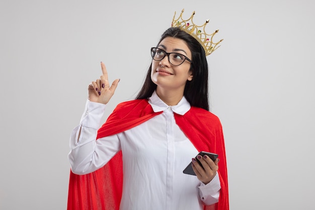 Foto grátis jovem supermulher satisfeita de óculos e coroa segurando o celular, olhando e apontando para cima, isolado na parede branca