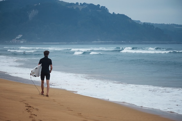 Jovem surfista com roupa de neoprene caminha pela praia com sua prancha de surf branca olhando para as ondas