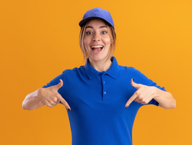 Jovem, surpresa, bonita, entregadora de uniforme, apontando para baixo com as duas mãos em laranja