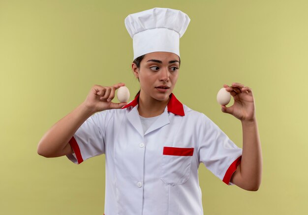Jovem surpresa caucasiana cozinheira com uniforme de chef segurando ovos isolados em uma parede verde com espaço de cópia