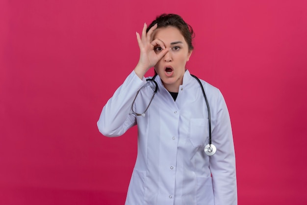 Jovem, surpresa, médica, vestindo bata médica e estetoscópio, mostrando o gesto do binóculo com uma das mãos sobre fundo vermelho isolado