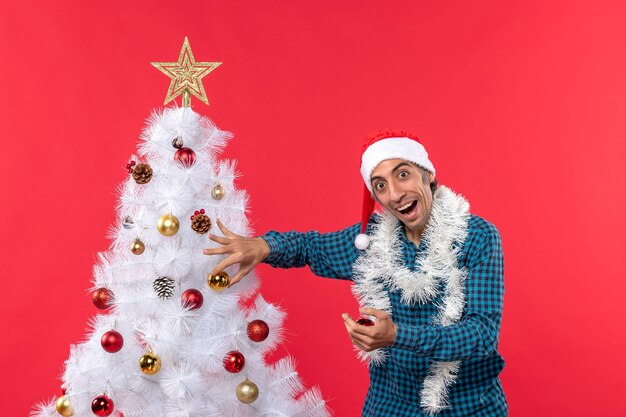 Jovem surpreso com chapéu de papai noel em uma camisa azul listrada e decorando uma árvore de natal surpreendentemente vermelha