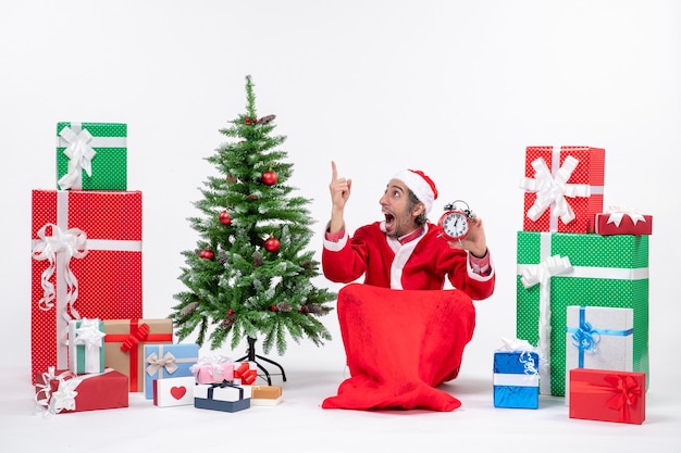 Jovem surpreso comemorar o ano novo ou o feriado de natal sentado no chão segurando o relógio perto dos presentes e decorando a árvore de natal no fundo branco