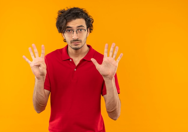 Jovem surpreso de camisa vermelha com óculos ópticos, gestos nove com dedos isolados na parede laranja