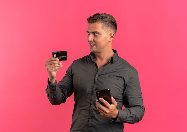 Foto grátis jovem surpreso loiro bonito segurando o telefone e olhando para o cartão de crédito