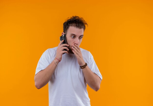 Jovem surpreso vestindo uma camiseta branca falando ao telefone e colocando a mão na boca em um fundo laranja isolado