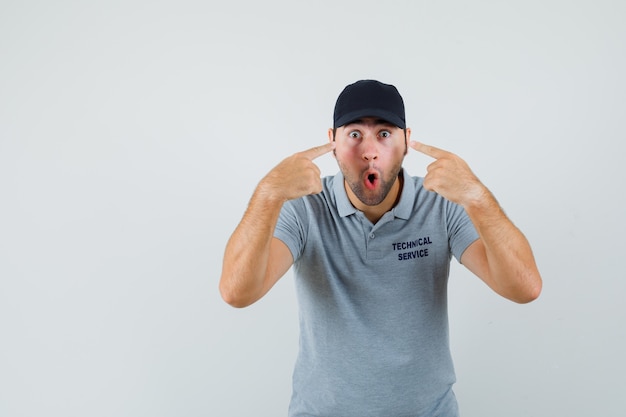 Jovem técnico apontando para os olhos em uniforme cinza e parecendo surpreso.