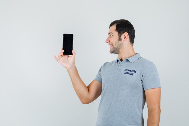 Jovem técnico de uniforme cinza, segurando o smartphone na mão e sorrindo enquanto olha para ele e olha a vista frontal otimista.