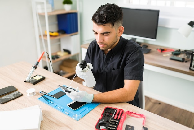 Foto grátis jovem técnico masculino usando ar comprimido para limpar dentro de um smartphone sujo na oficina. homem latino com luvas limpando um celular