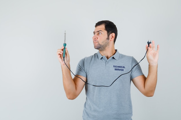 Jovem técnico olhando para a chave de fenda segurando sua mão em uniforme cinza e olhando com foco.