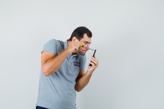 Jovem técnico segurando a chave de fenda e abrindo a parte de trás de seu telefone em uniforme cinza e olhando com foco.