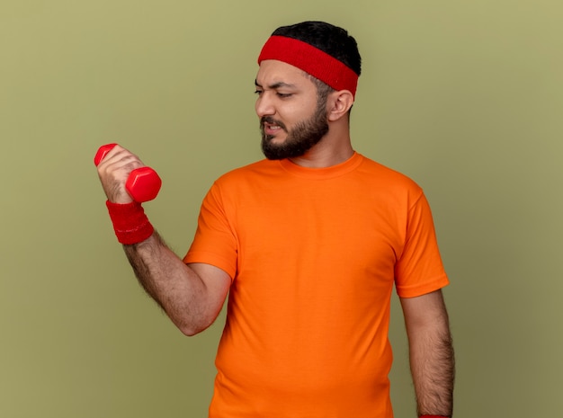 Foto grátis jovem tenso e esportivo usando bandana e pulseira, fazendo exercícios com halteres isolado em fundo verde oliva