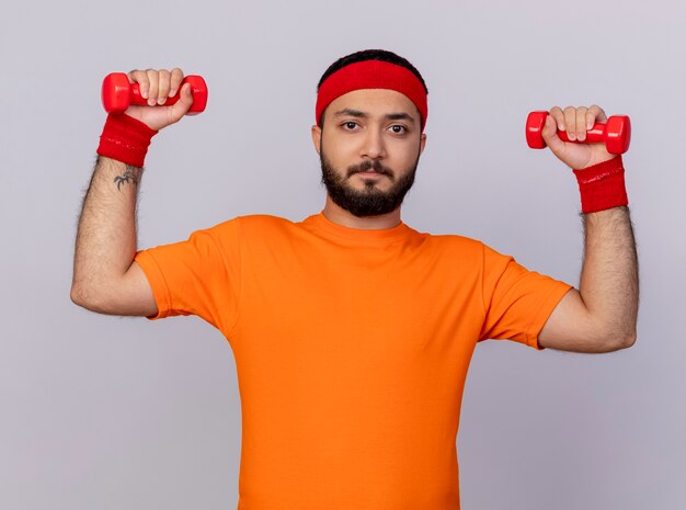 Jovem tenso e esportivo usando bandana e pulseira, levantando halteres isolados no fundo branco