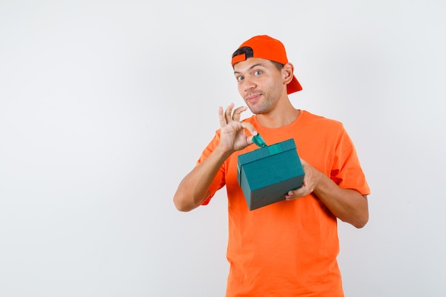 Jovem tentando abrir uma caixa de presente com uma camiseta laranja e boné e parecendo curioso